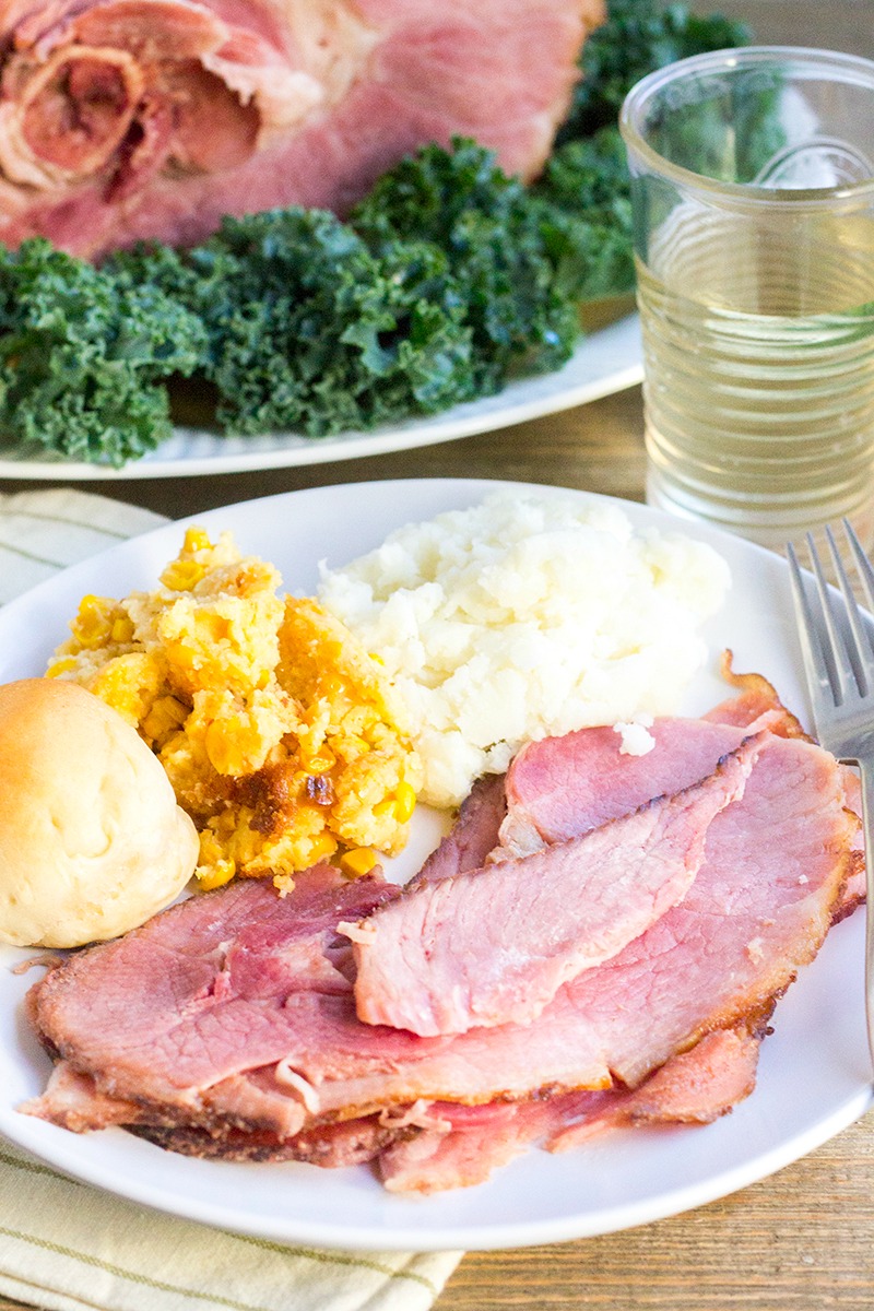 slices of ham on a plate with potatoes, corn, and a roll