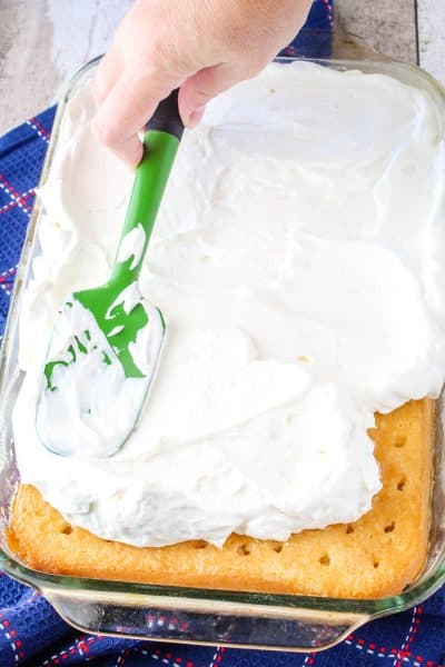 whipped cream being spread over a coconut poke cake