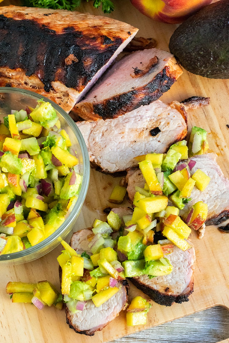 grilled pork tenderloin slices on a cutting board topped with avocado peach salsa