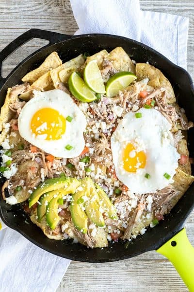 skillet of pork chilaquiles recipe garnished with queso fresco, onion, tomato, avocado, and lime