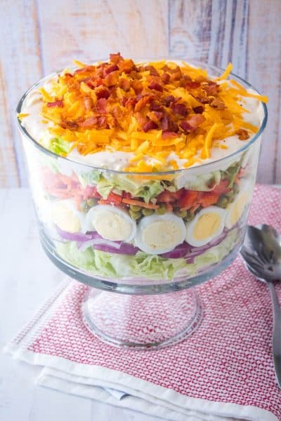seven layer salad in a trifle bowl with serving spoons