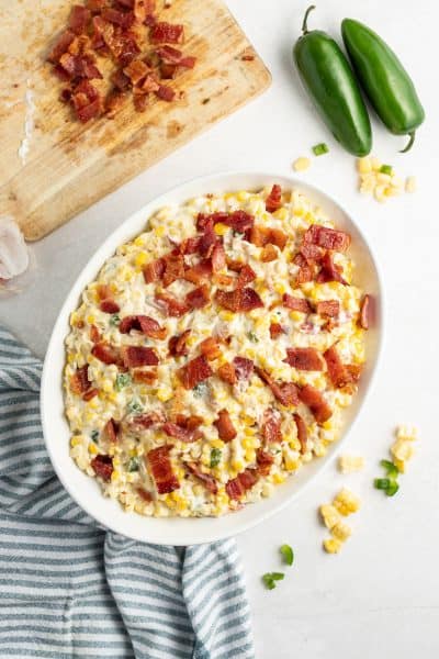 chopped bacon on a cutting board next to two jalapenos and a casserole dish of creamed corn