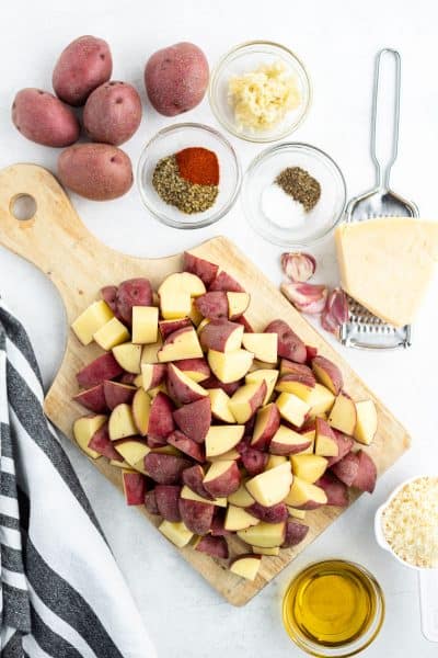 cubed red potatoes on a cutting board with bowls of oil, spices, and Parmesan cheese