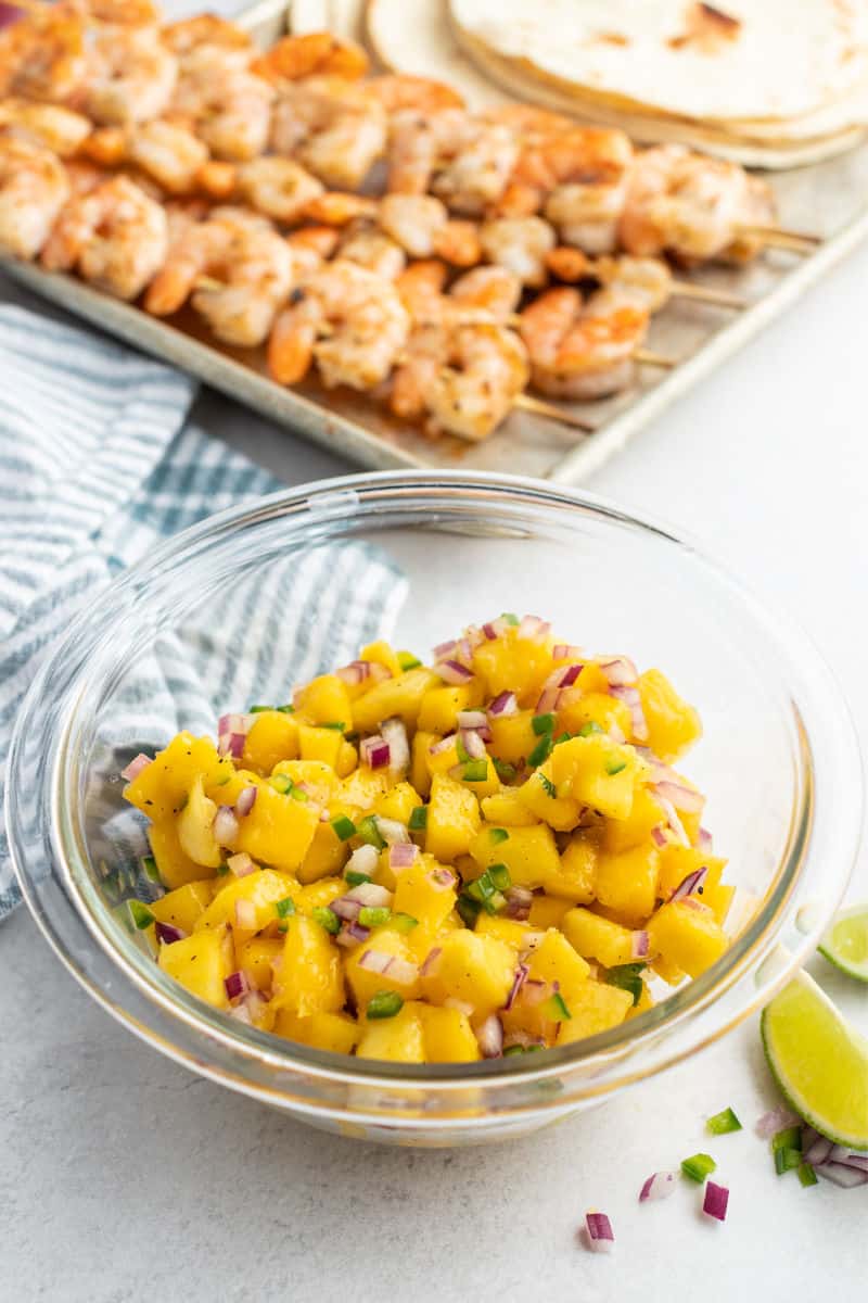 mango salsa in a glass bowl in front of cooked shrimp skewers