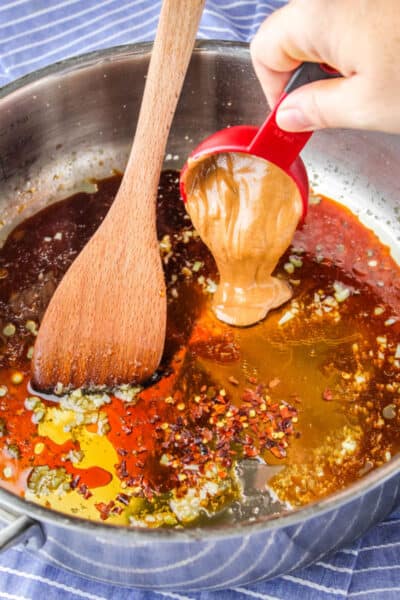 honey almond butter being added to a skillet with cooked garlic, ginger, soy sauce, and other peanut sauce ingredients