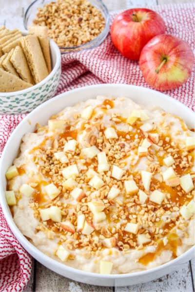 caramel apple cheesecake dip in a serving bowl next to graham crackers, chopped nuts, and gala apples