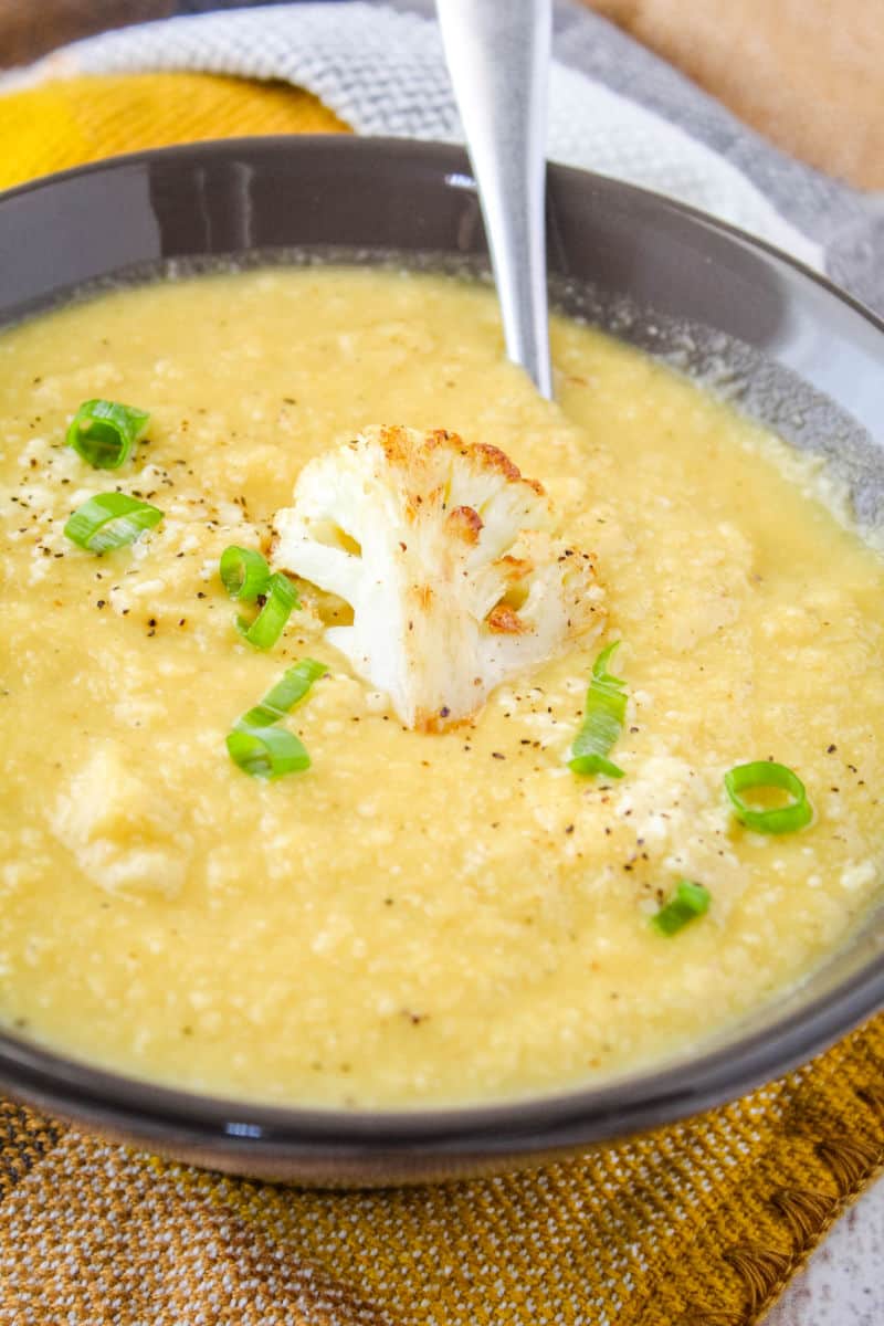 close up on roasted garlic & cauliflower soup in a bowl with a spoon, piece of cauliflower, green onion, and pepper on top