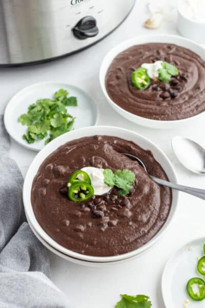 two bowls of black bean soup with a plate of cilantro and a plate of jalapeno slices nearby
