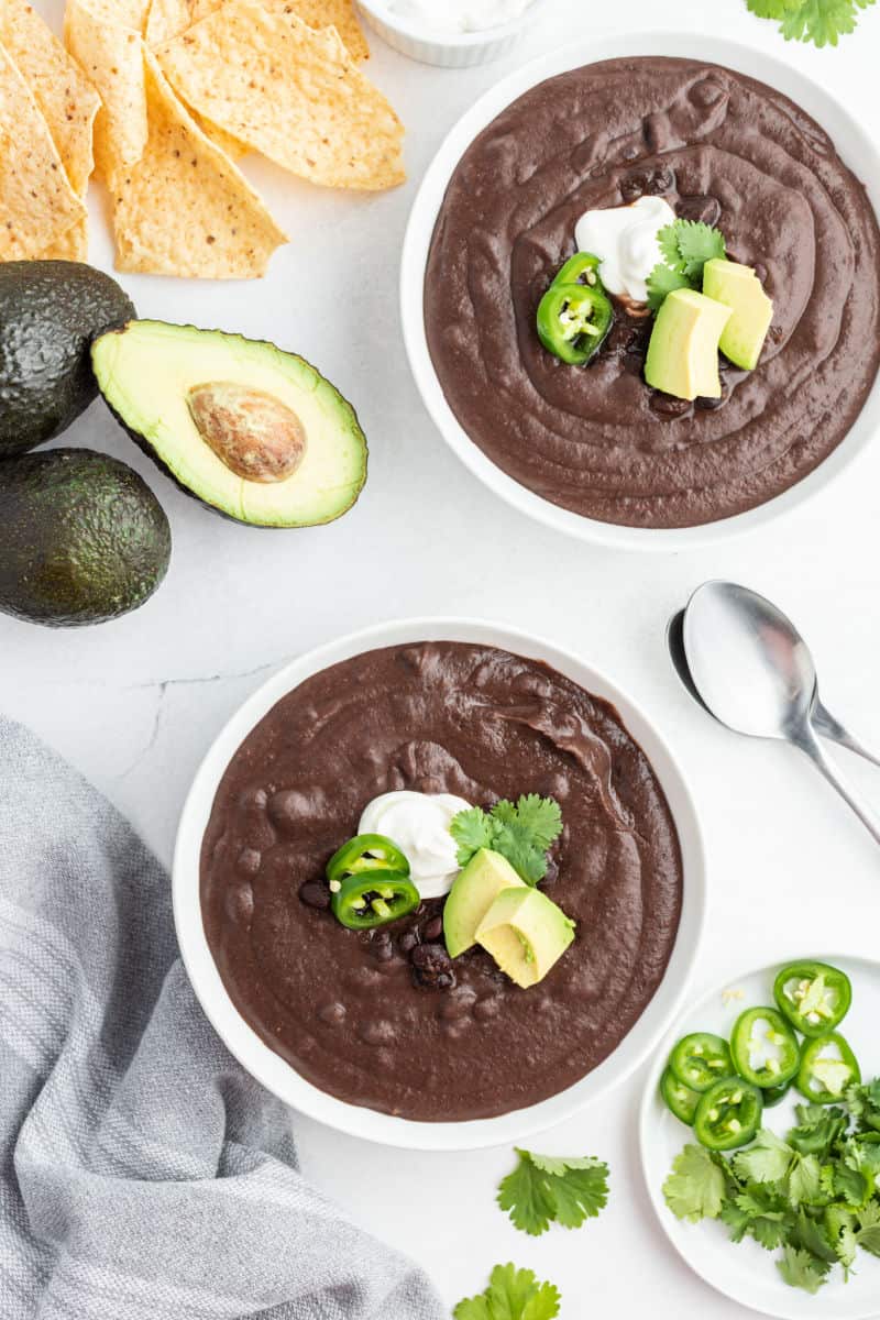 two bowls of slow cooker black bean soup next to an avocado cut in half and tortilla chips