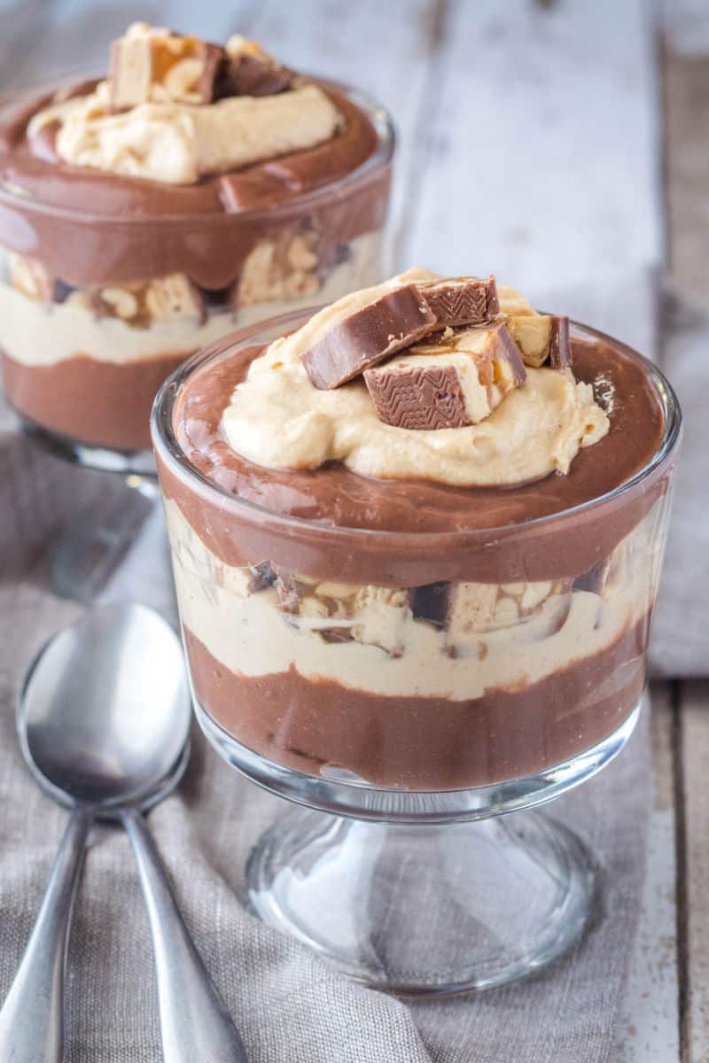 two snickers chocolate parfaits in dessert cups with spoons next to them