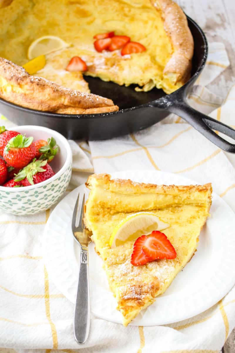slice of german pancake on a small plate with powdered sugar, lemon wedge, and sliced strawberries on top