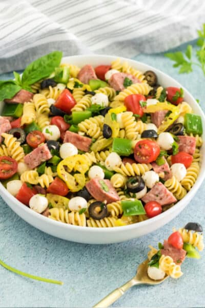 side view of anitpasto pasta salad in a white serving bowl next to a fork with pasta salad on it