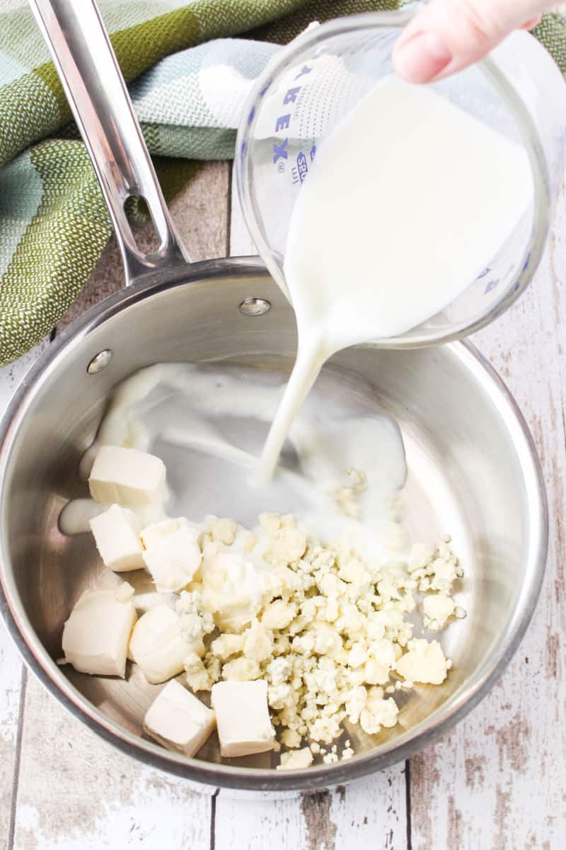 milk being poured into a saucepan with cream cheese cubes and blue cheese crumbles