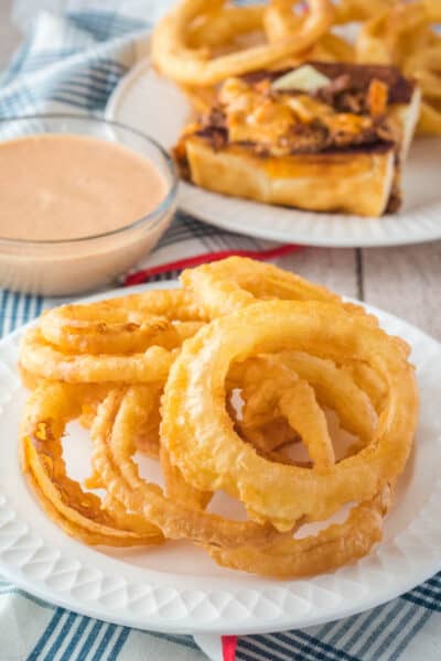 onion rings on a plate next to a bowl of fry sauce with a chili dog in the background