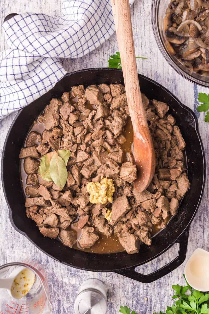 cooked beef with beef broth, bay leaves, and mustard in a skillet