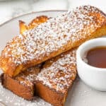 square image of three french toast sticks on a plate next to a cup of syrup