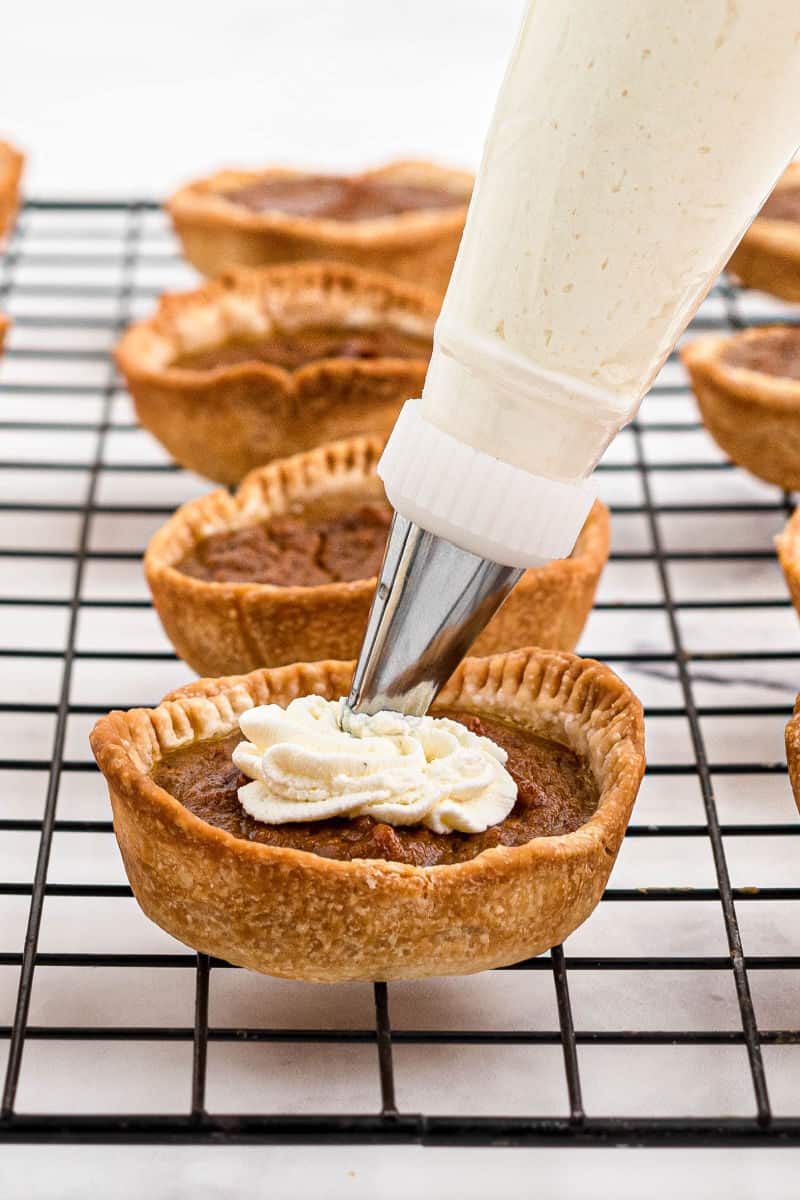 whipped cream being piped onto mini pumpkin pies on a wire rack