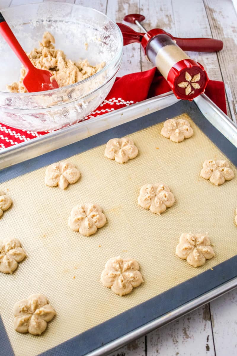 spritz cookies on a baking sheet next to a bowl of cookie dough and a cookie press