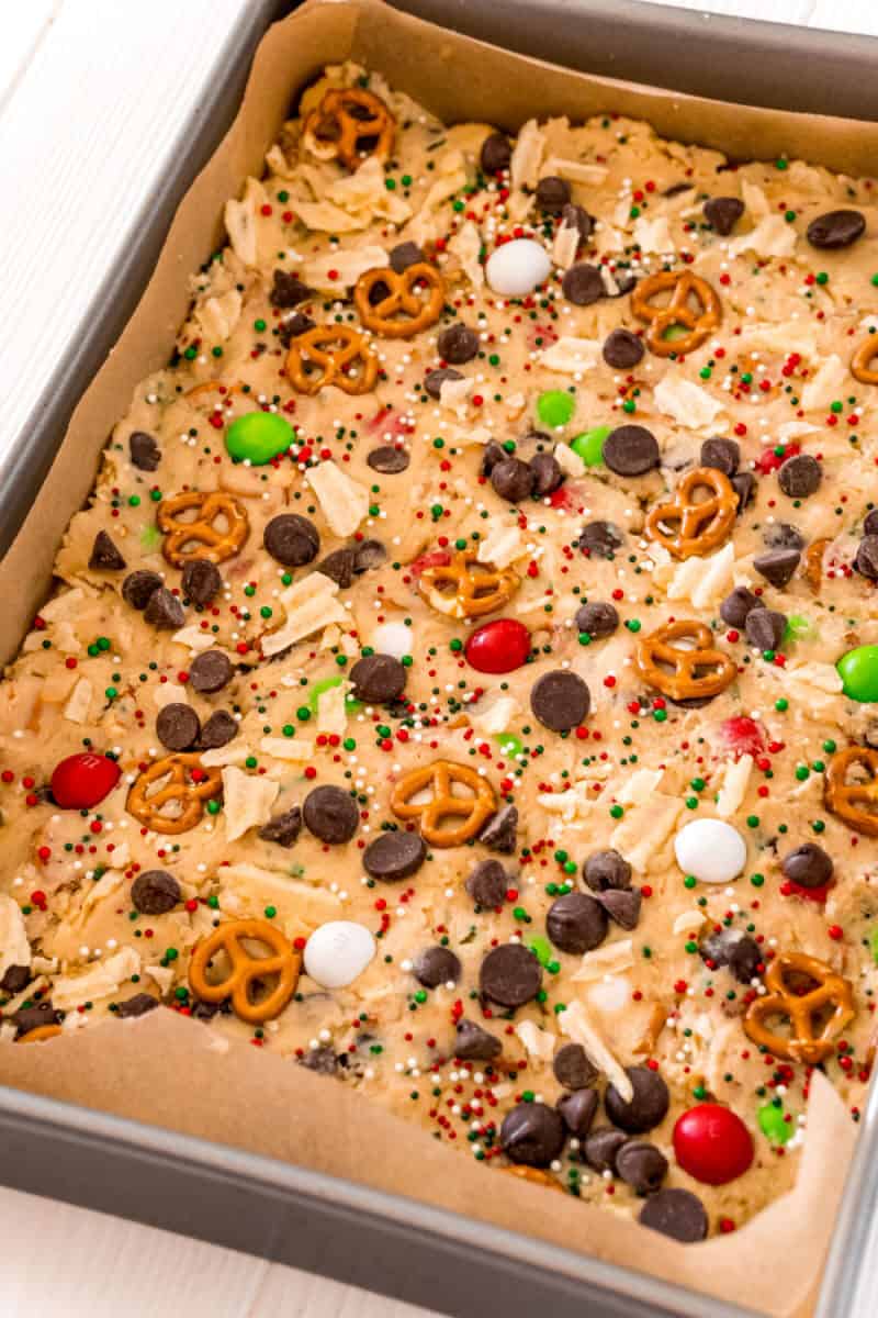 cookie bar dough in a bakign dish topped with christmas M&Ms, potato chips, pretzels, chocolate chips & sprinkles