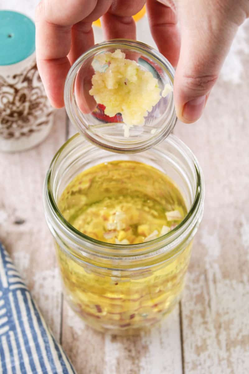 garlic being added to a mason jar with olive oil, vinegar, and shallots