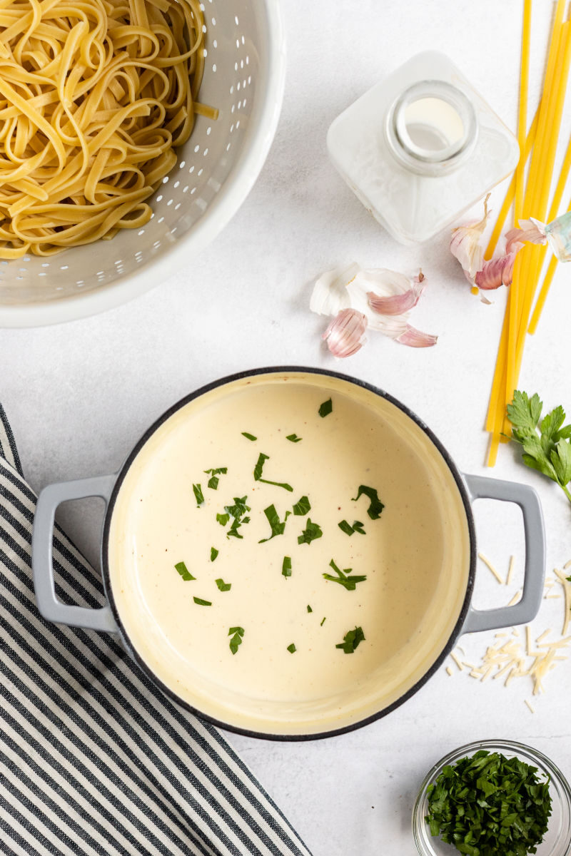 finished alfredo sauce with chopped parsley in a pot next to a colander with cooked fettuccine