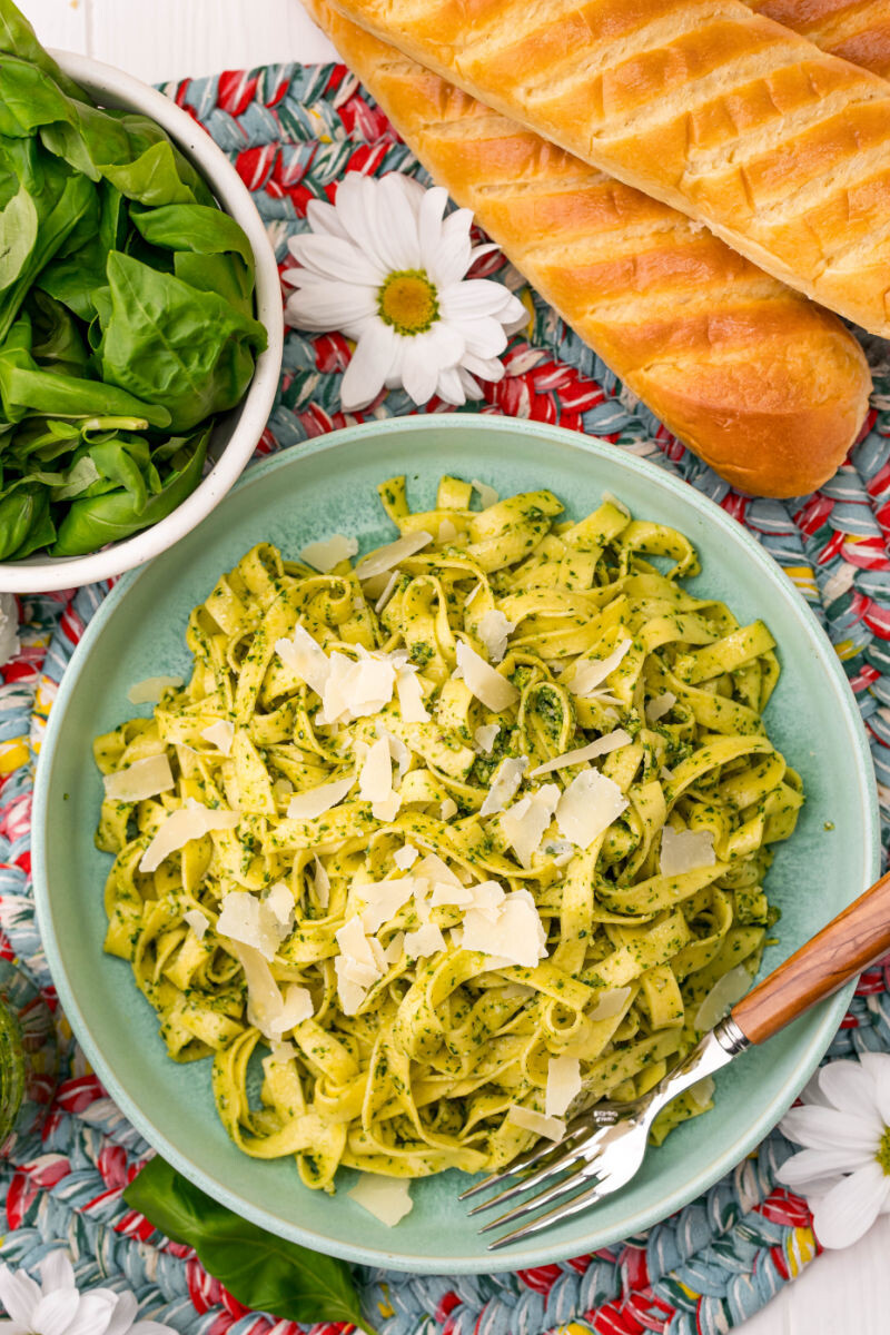 plate of pesto pasta topped with parmesan next to bread and a bowl or basil leaves