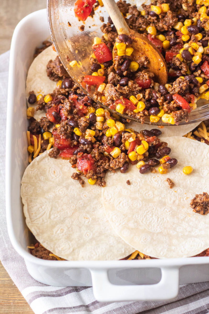 taco meat and vggie mixture being poured on top of corn tortillas layered in a baking dish