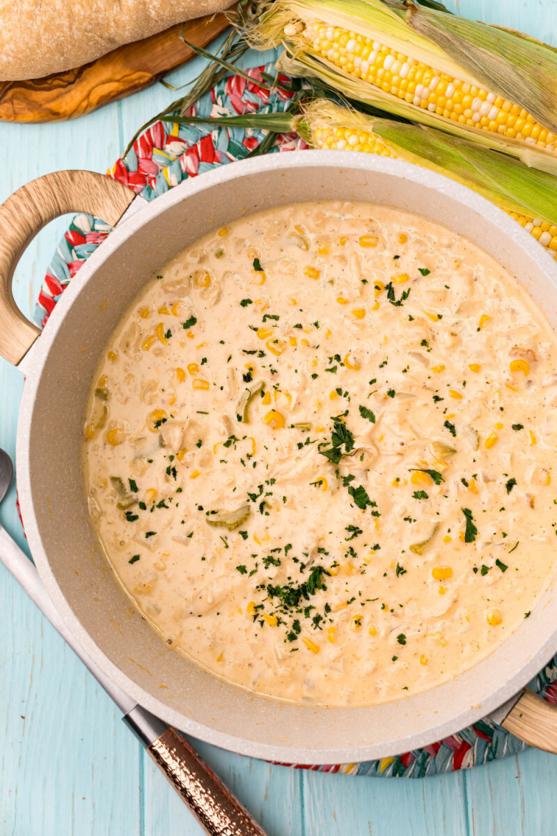 chicken corn chowder in a pot topped with chopped parsley next to ears of corn