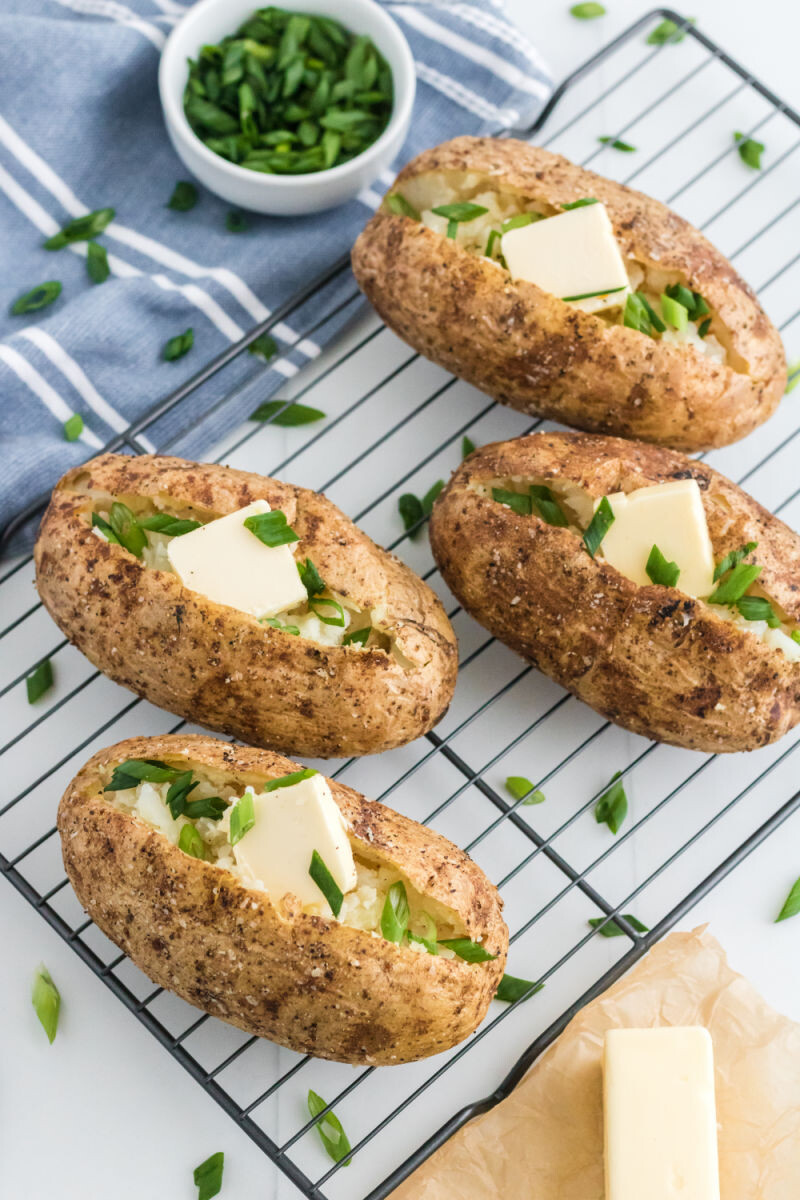four baked potatoes on a wire rack topped with butter and sliced green onions