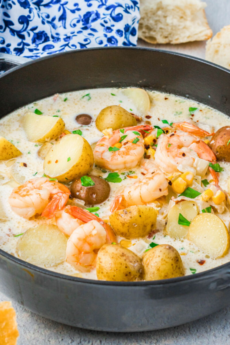 side view of shrimp and corn chowder with potatoes in a pot with hunks of bread in the background