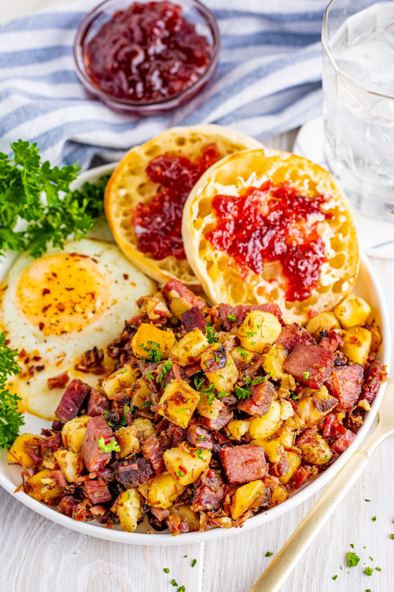 corned beef hash on a plate with sunny side up eggs and English muffins with jelly
