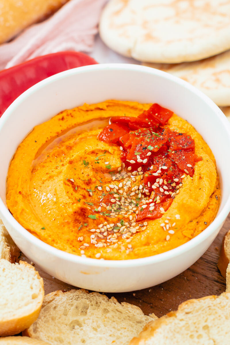 roasted red pepper hummus in a bowl topped with sesame seds, herbs, and roasted red peppers