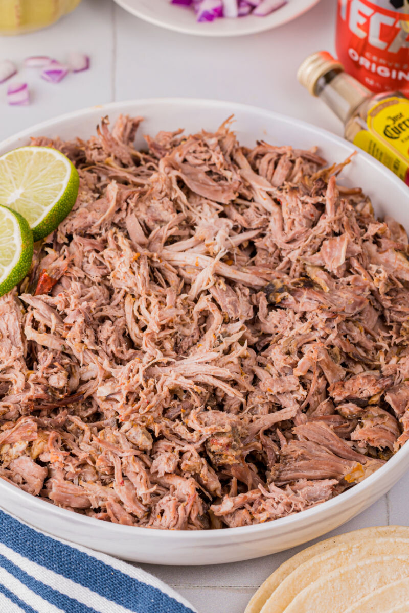 bowl of slow cooker carnitas with two lime slices for garnish next to a bottle of tequila and can of Mexican beer