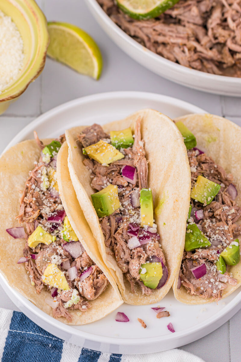 slow cooker carnitas tacos with avocado, red onion, and cojita cheese on top
