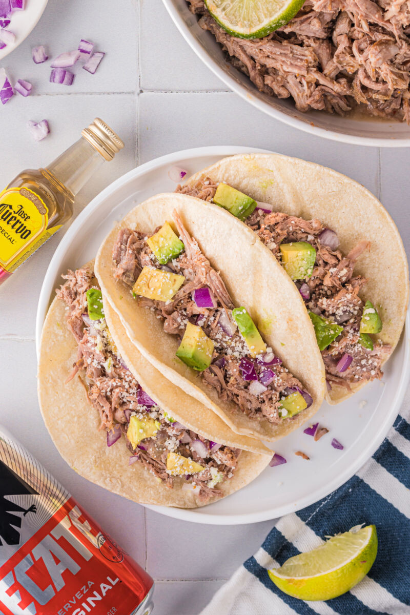 looking down at three slow cooker carnitas tacos with avocado, red onion, and cojita