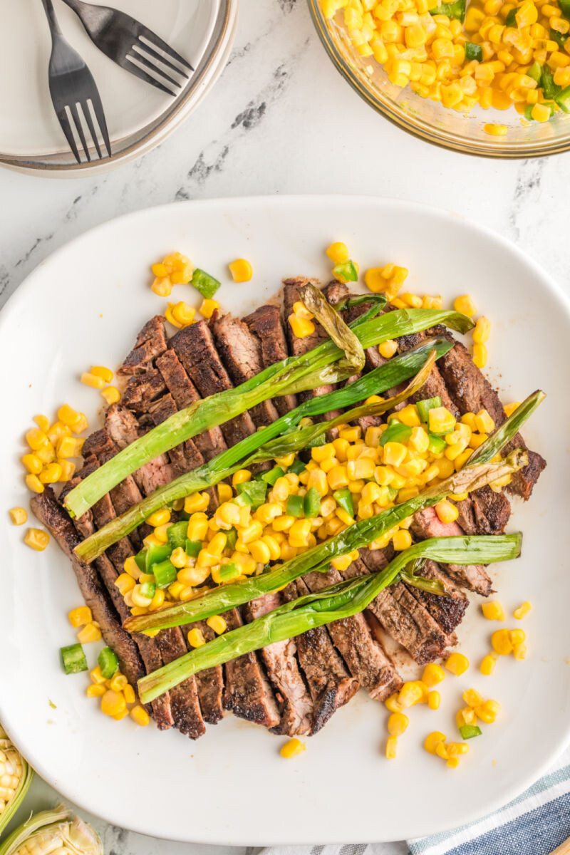 sliced seared flank steak on a platter topped with wilted green onions and corn salsa