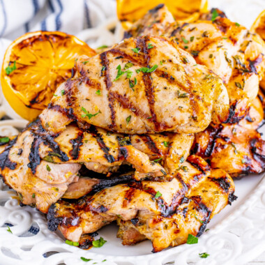 square image of lemon garlic grilled chicken thighs piled up on a serving platter with grilled lemons