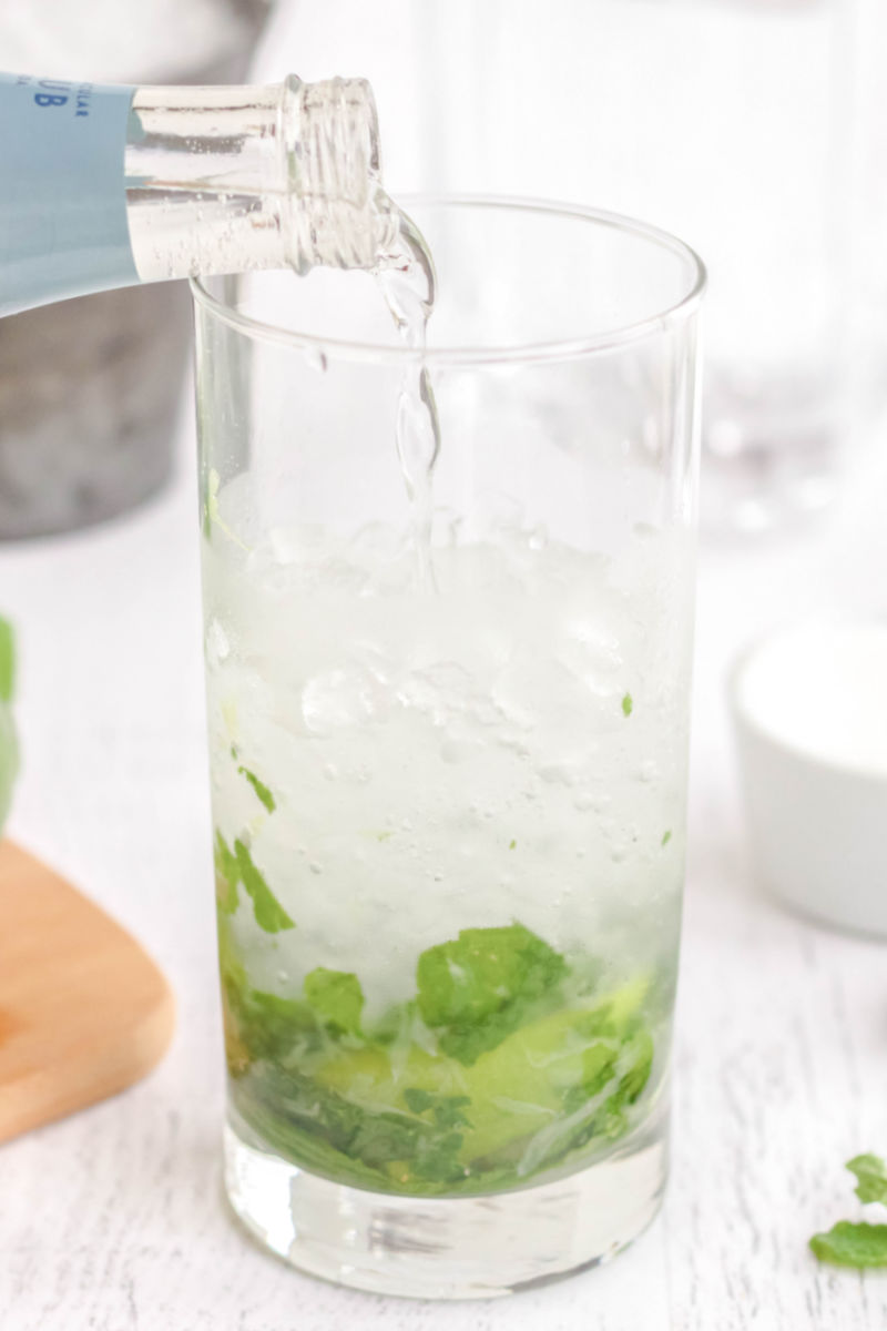 soda water being poured into a glass with ice, rum, mint, lime, and simple syrup