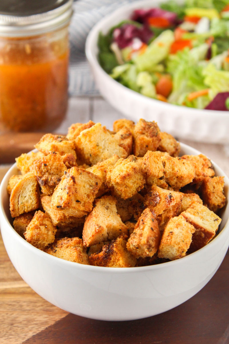 homemade croutons in a bowl next to a salad and jar of dressing