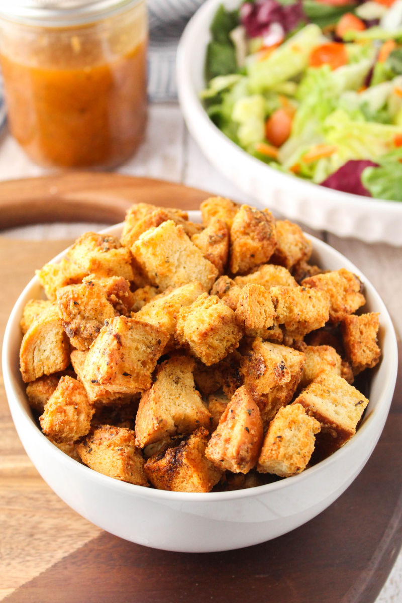 angled view of homemade garlic parmesan croutons in a bowl next to a salad and dressing