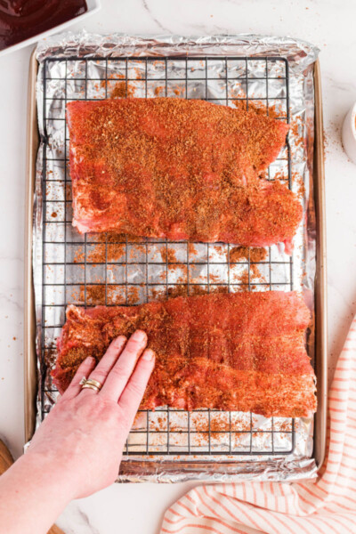 dry rub for ribs being rubbed into half rack of ribs on a rack over a baking sheet