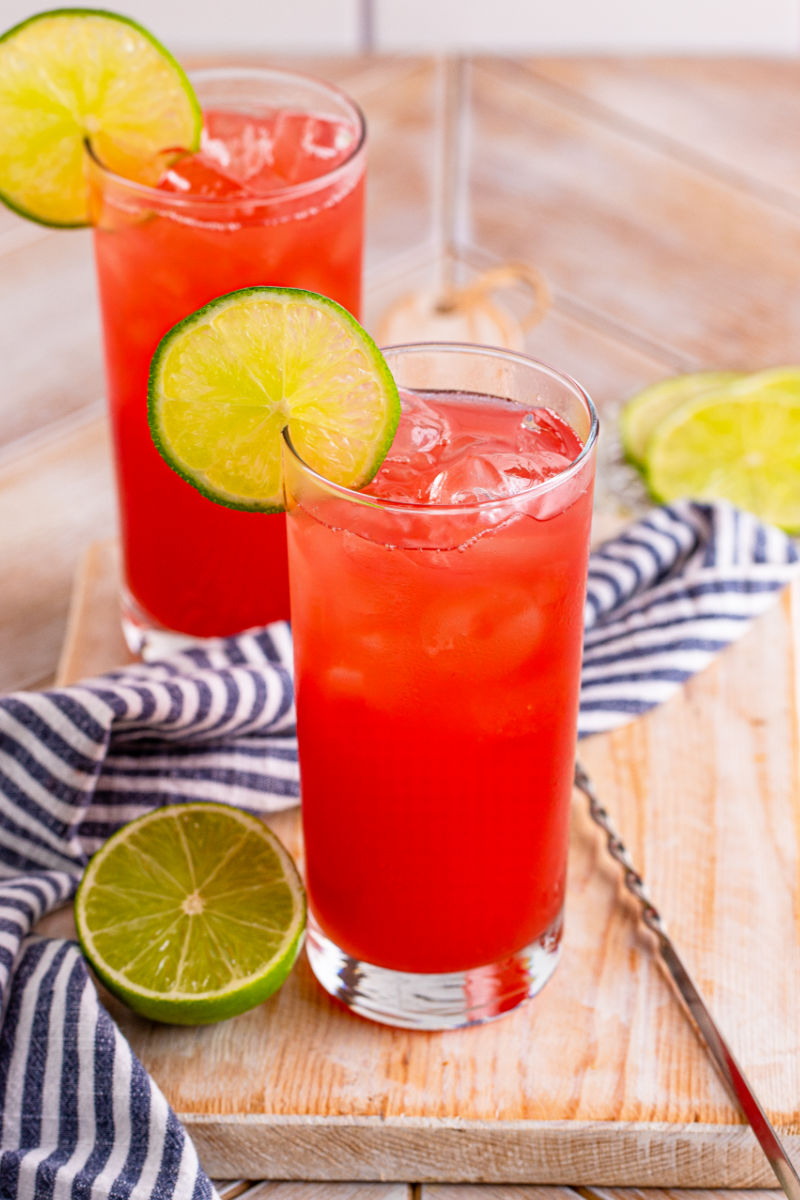 two bay breeze cocktails on a wooden board next to half of a lime and a bar spoon