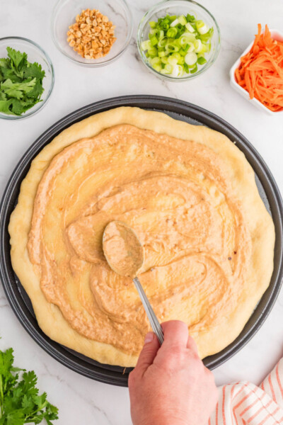 thai peanut sauce being spread over a par-baked pizza crust with a spoon