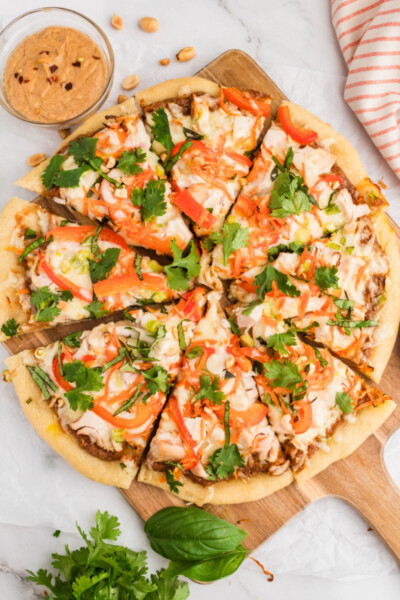 thai chicken pizza topped with basil, cilantro, and peanuts and cut into slices on a cutting board