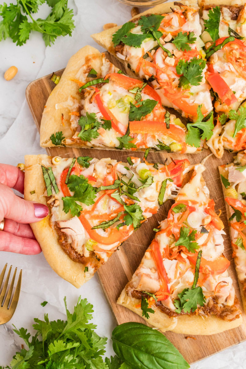 hand taking a slice of thai chicken pizza from the serving board
