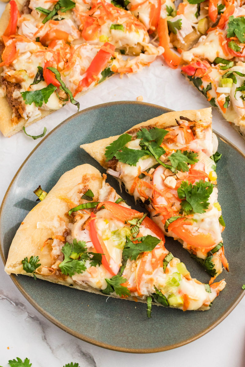 two slices of thai chicken pizza on a small plate next to the pizza